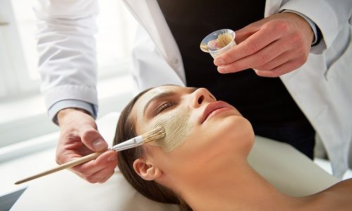 Male cosmetologist putting mask on young female face with a brush during cosmetic procedure.